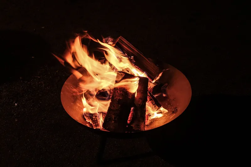 Feuerschale mit brennenden Holzscheiten und hellen Flammen im Dunkeln, im Freien auf dunklem Boden