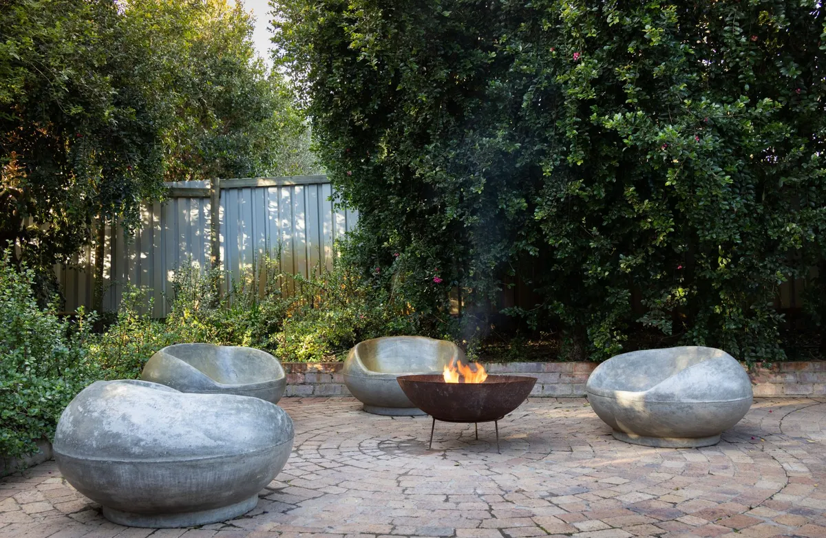 Brennende Feuerschale aus Metall auf gepflastertem Boden, umgeben von steinernen Sitzschalen und Gartenhecken